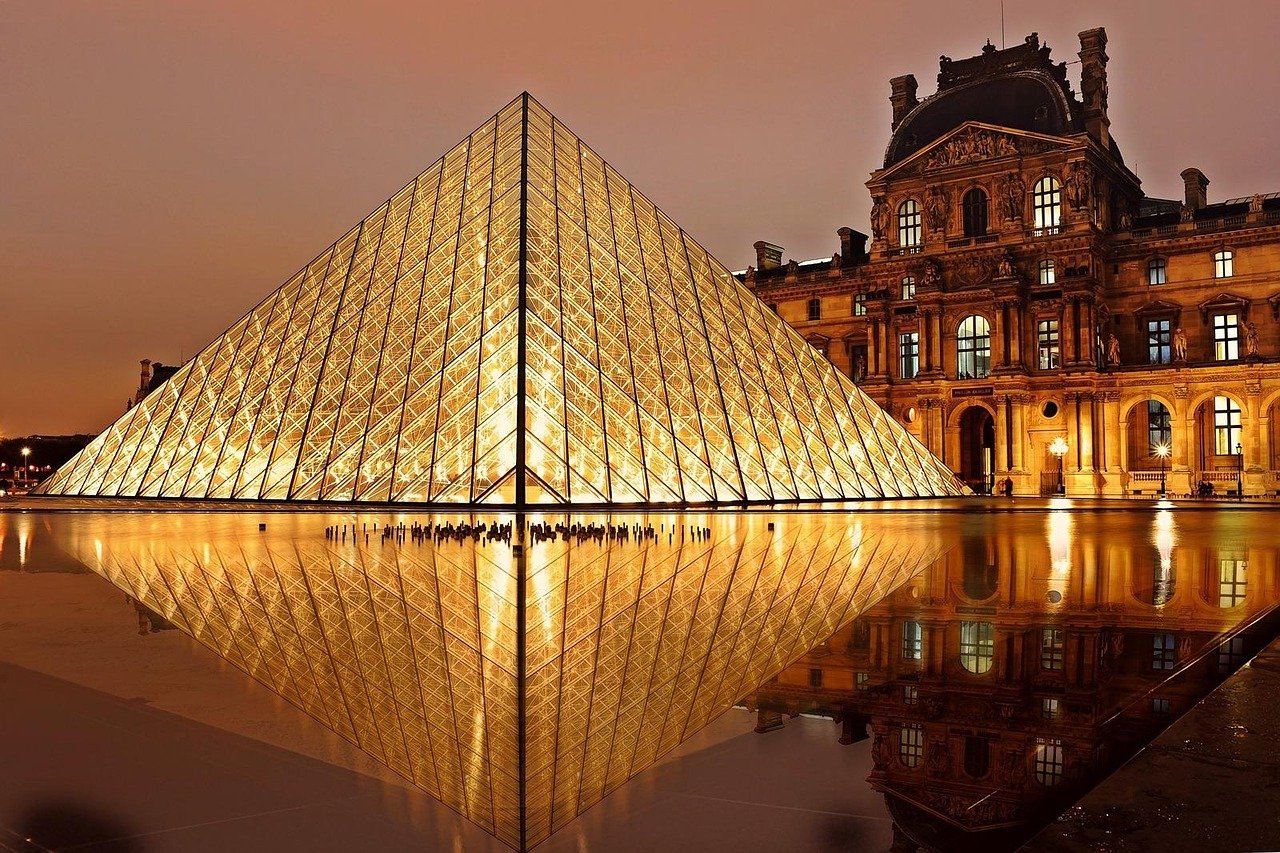 Museu do Louvre: Ingressos, Como Evitar Filas e o Que Ver 1 Pirâmide de vidro do Louvre iluminada ao entardecer, refletida na água